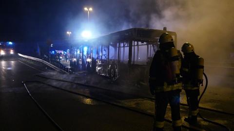 Ein ausgebranntes Wrack eines Linienbusses auf einer Bundesstraße im Dunkeln, daneben Feuerwehrleute.