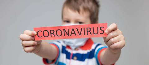 Ein Kind hält Schild mit der Aufschrift "Coronavirus" in den Händen und streckt es nach vorne in Richtung Kamera.