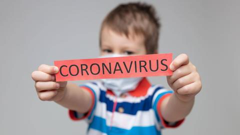 Ein Kind hält Schild mit der Aufschrift "Coronavirus" in den Händen und streckt es nach vorne in Richtung Kamera.