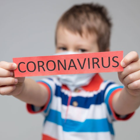 Ein Kind hält Schild mit der Aufschrift "Coronavirus" in den Händen und streckt es nach vorne in Richtung Kamera.