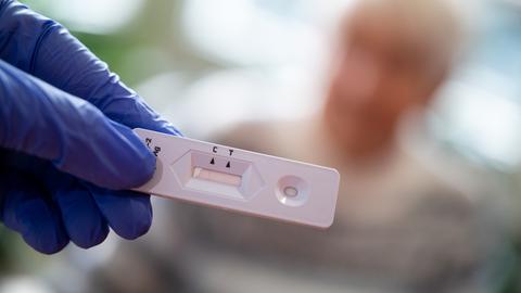 Eine Hand, die einen Antigen-Corona-Schnelltest in der Hand hält, im Hintergrund unscharf ein älterer Mensch.