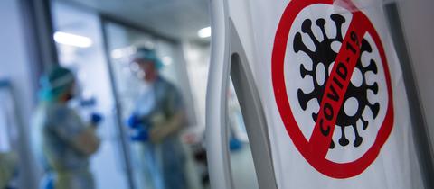 Ein Symbol - im Bildvordergrund ein Virus-Icon in einem roten durchgestrichenen Kreis - an der Tür weist auf den Covid-Bereich der Klinik hin. Im Hintergrund unscharf zwei Ärzte im Gespräch.