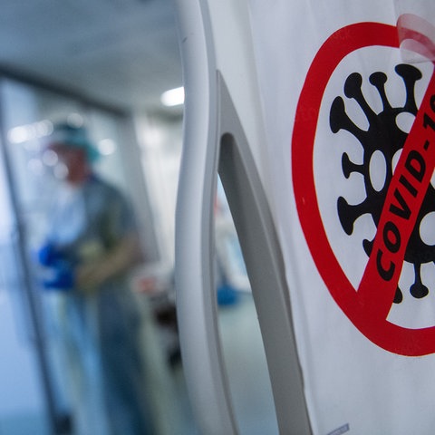 Ein Symbol - im Bildvordergrund ein Virus-Icon in einem roten durchgestrichenen Kreis - an der Tür weist auf den Covid-Bereich der Klinik hin. Im Hintergrund unscharf zwei Ärzte im Gespräch.