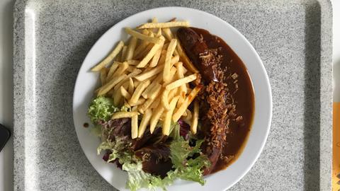 Ein Teller mit Currywurst, Pommes frites und einer kleinen Salatbeilage steht auf einem Tablet in einer Kantine.