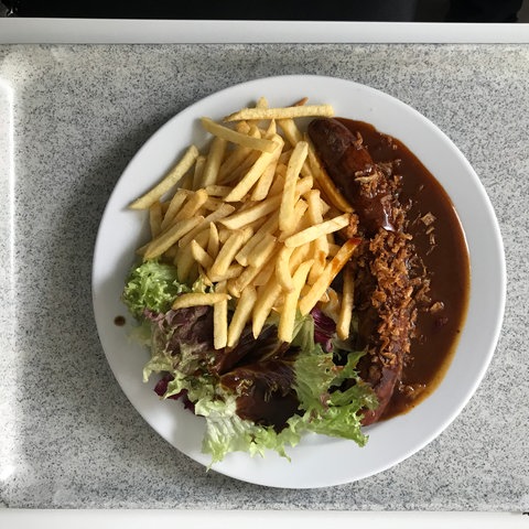 Ein Teller mit Currywurst, Pommes frites und einer kleinen Salatbeilage steht auf einem Tablet in einer Kantine.