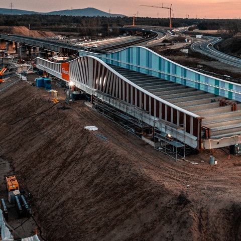 Großbaustelle von oben in der Dämmerung. Ein Teil einer Autobahnbrücke wird über eine Autobahn, die im Bau ist, geschoben.