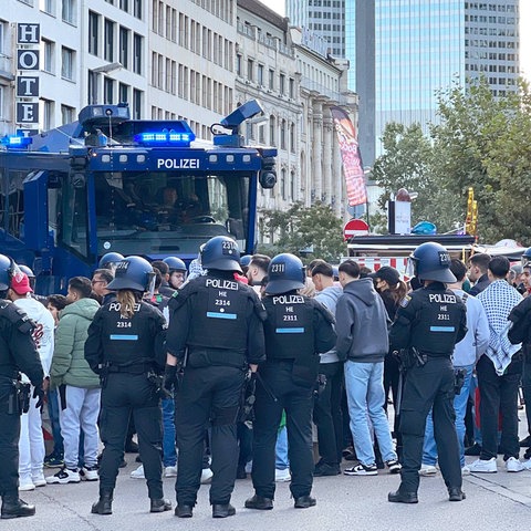 Viele Polizisten stehen um eine kleine Gruppe von Menschen im öffentlichen Raum. Daneben steht ein Wasserwerfer.