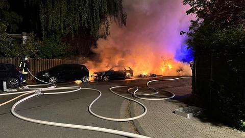 Drei Autos stehen in Flammen, daneben ein Feuerwehrmann, Schläuche liegen auf dem Boden.