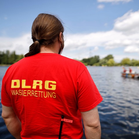 Ein Rettungsschwimmer steht mit dem Rücken zur Kamera vor einem See, DLRG steht auf seinem T-Shirt.
