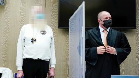 Der Angeklagte steht im Verhandlungssaal neben seinem Anwalt an einem Tisch. Zwischen ihnen eine Plexiglasscheibe zum Infektionsschutz.