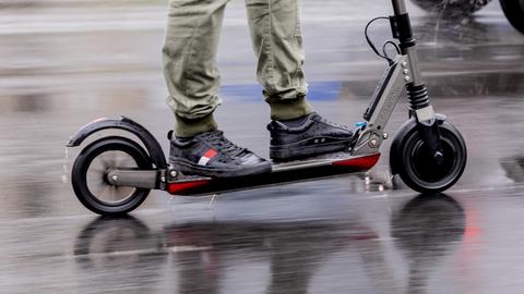 Mensch auf E-Scooter in Fahrt auf einer nassen Straße. Daneben ein Auto.