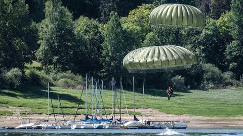 Fallschirmspringer in der Luft über einem See, an dessen Ufer Boote liegen.