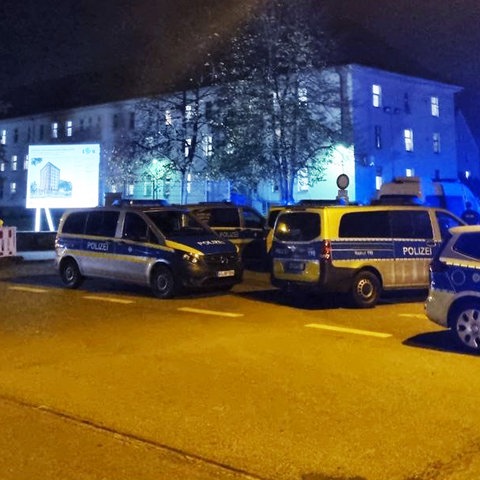 Viele Polizeiautos im Einsatz auf einer Straße vor einem erleuchteten Flüchtlingsheim.