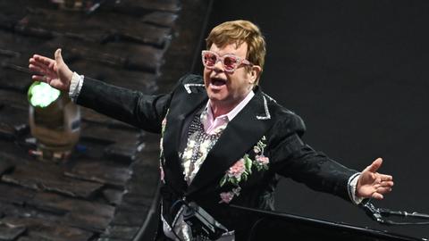 Der Musiker Elton John in einem schwarzen Anzug mit rosa Brille singt auf einer Bühne und breitet dabei die Arme aus.