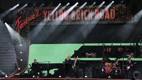 Auf einer Bühne spielt eine Band vor einem schwarzen Hintergrund mit der Aufschrift "Farewell Yellow Brick Road".