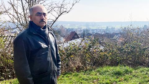 Ein Mann steht auf einer Wiese vor einem landschaftlichen Panorama und guckt traurig in die Kamera.