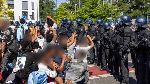 Menschen mit verpixelten Gesichtern, Fäusten in der Luft, manche am Boden, vor einer Polizeikette.
