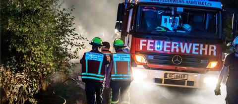 Auf einem Waldweg nähern sich in der Nacht mehrere Feuerwehrmänner in Ausrüstung und mit aufgeklapptem Visier einem Feuerwehrwagen, dessen Strahler den Weg ausleuchten.