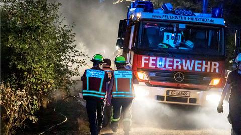 Auf einem Waldweg nähern sich in der Nacht mehrere Feuerwehrmänner in Ausrüstung und mit aufgeklapptem Visier einem Feuerwehrwagen, dessen Strahler den Weg ausleuchten.