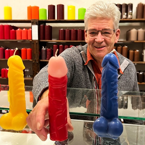 Ein Mann greift mit der Hand nach Kerzen in Penisform im Vordergrund 
