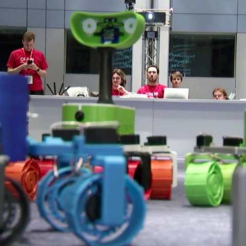 Im Bildvordergrund grpß bunte Plastikteile, im Hintergrund Menschen vor Bildschirmen an einem Desk sitzend.