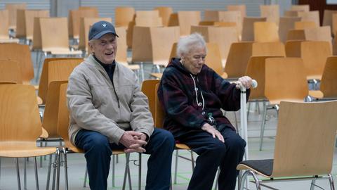 Zwei ältere Anwohner sitzen in einer Sporthalle in Hanau auf einem Holzstuhl.