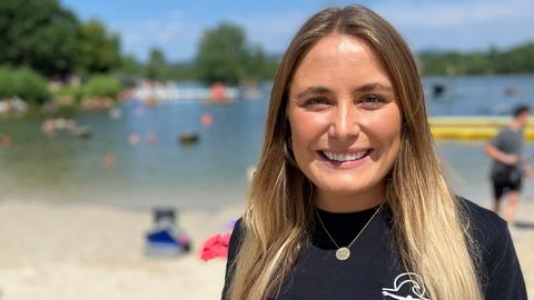 Eine Frau in einem schwarzen T-Shirt steht am Strand vor einem Badesee.