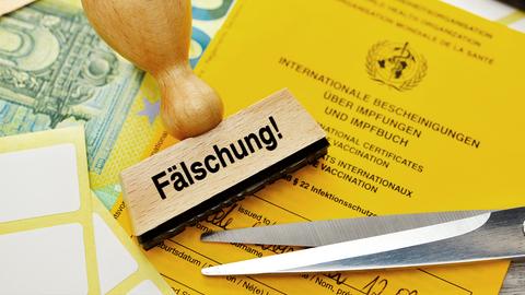 Ein Impfpass, eine Stempel mit der Aufschrift "Fälschung", eine Schere und Geld liegen zusammen und übereinander auf einem Tisch.