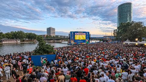 Fans stehen vor dem Big Screen auf der Fanzone, im Hintergrund ist der Main zu sehen. 