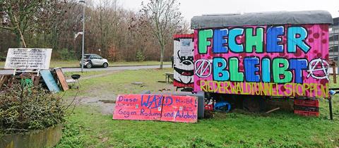 Auf einer Grünfläche steht ein Bauwagen, auf dem steht "Fecher bleibt, Riederwaldtunnel stoppen"