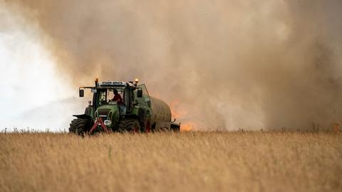 Ein Traktor fährt über ein Getreidefeld, dahinter lodern Flammen und es steigt Rauch auf.