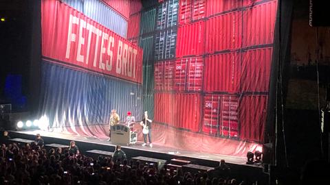 Auf einer Bühne in einer Konzerthalle stehen drei Männer, dahinter ein Vorhang mit der Aufschrift "Fettes Brot"