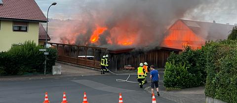 Eine Werkstatt steht lichterloh in Flammen - davor Feuerwehrleute. 