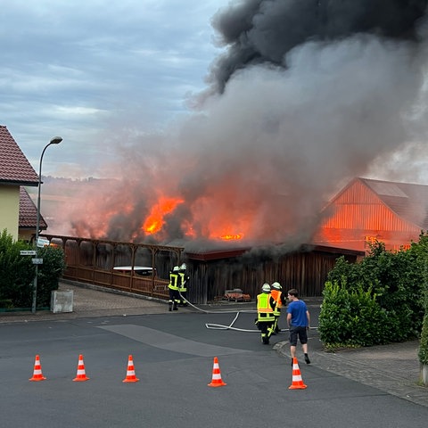 Eine Werkstatt steht lichterloh in Flammen - davor Feuerwehrleute. 