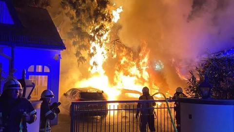Hinter einem Metallzaun brennt es meterhoch - mehrere Autos stehen in Flammen. Die Flammen züngeln bis hoch zu einem Baum.