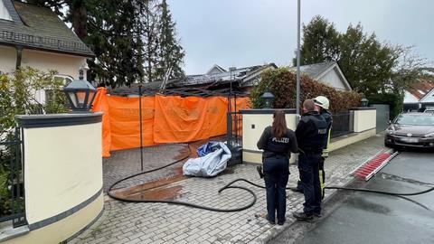 Polizeibeamte stehen bei Tageslicht vor einer Einfahrt. Das Dach einer Garage ist zerstört. Als Sichtschutz wurde vor den zerstörten Autos eine Plane angebracht.