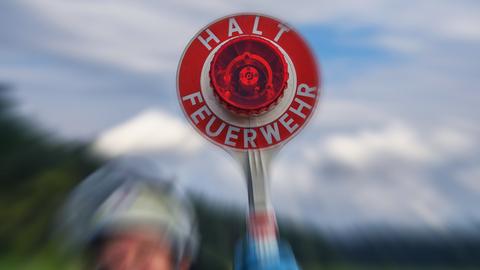 Im Bildvordergrund eine Kelle mit der Aufschrift "Halt Feuerwehr". Im Hintergrund - unscharf - der Kopf der Einsatzkraft, welche die Kelle hält.