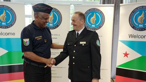 Feuerwehrchef Colonel Moussa Mahamoud und Lothar Hügin, Präsident der international fire and rescue confederation (IFCR) geben sich die Hand, im Hintergrund sind die Flaggen der beiden Länder zu sehen: Deutschland und Djibouti