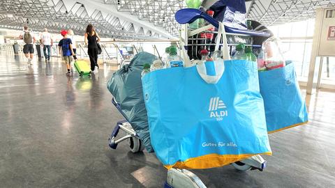Ein Gepäckwagen mit großen Tüten der Marke Aldi und Pfandflaschen im Bildvordergrund. Im Bildhintergrund Reisende von hinten mit Gepäck in einer großen Halle.