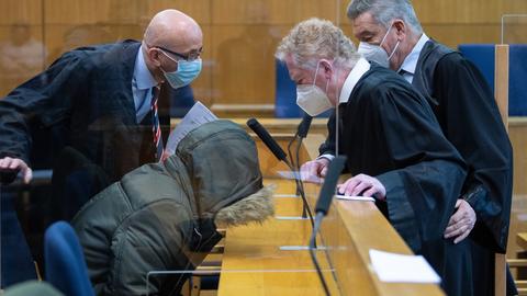 Der Angeklagte sitzt in einem Parka mit hochgezogener Kapuze am Tisch mit gebeugtem Kopf. Daneben sprechen seine Verteidiger miteinander.
