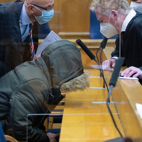 Der Angeklagte sitzt in einem Parka mit hochgezogener Kapuze am Tisch mit gebeugtem Kopf. Daneben sprechen seine Verteidiger miteinander.