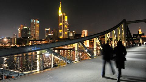 Zwei Menschen gehen auf einer Fußgängerbrücke (Bildvordergrund). Im Bildhintergund leuchten die Bankentürme der Skyline in de Nacht.