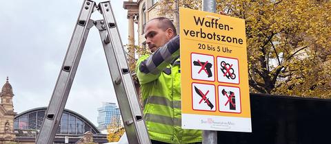 Ein Mann in neonfarbener Arbeitskleidung steht auf einer Leiter und hängt ein Schild mit der Aufschrift "Waffenverbotszone" und entsprechenden Symbolen an einer Stange im Straßenraum auf. Im Hintergund ist der Hauptbahnhof Frankfurt zu sehen.