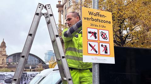 Ein Mann in neonfarbener Arbeitskleidung steht auf einer Leiter und hängt ein Schild mit der Aufschrift "Waffenverbotszone" und entsprechenden Symbolen an einer Stange im Straßenraum auf. Im Hintergund ist der Hauptbahnhof Frankfurt zu sehen.