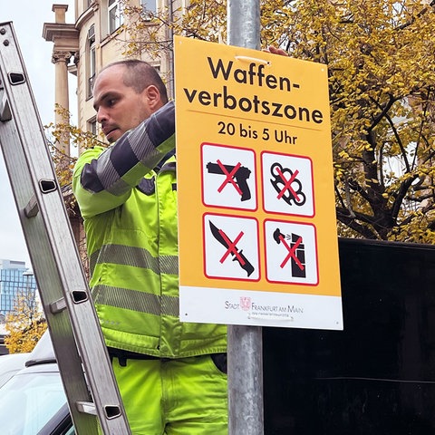 Ein Mann in neonfarbener Arbeitskleidung steht auf einer Leiter und hängt ein Schild mit der Aufschrift "Waffenverbotszone" und entsprechenden Symbolen an einer Stange im Straßenraum auf. Im Hintergund ist der Hauptbahnhof Frankfurt zu sehen.