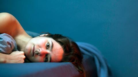 Eine Frau liegt in einem Bett mit blauer Bettwäsche. Es sind nur ihr Kopf und ihre unbekleideten Schultern zu sehen. Die Wände hinter dem Bett sind in derselben Farbe gestrichen. 