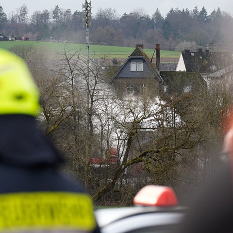 In der Ferne hinter Bäumen weißer Rauch, im Vordergrund sind Feuerwehrleute zu sehen.