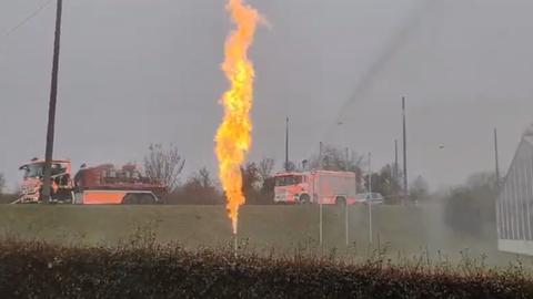 Gasflamme auf einem begrünten Gelände, Feuerwehrautos im Hintergrund. 