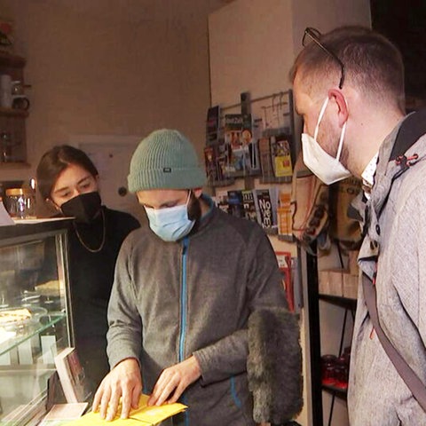 Drei Menschen stehen in einem Cafe am Tresen. Eine Person vergleicht zwei gelbe Impfpässe, die vor ihnen auf dem Tresen liegen.