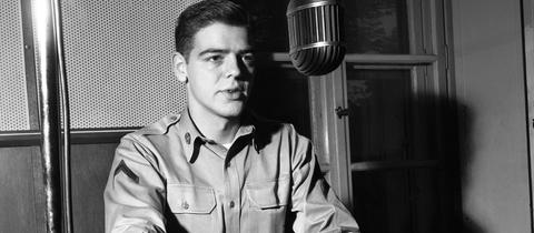 Radio-DJ Nick Clooney sitzt an einem Mikrofon und einem Plattenspieler im AFN-Studio in Frankfurt, Schwarz-weiß-Aufnahme aus den 1950er Jahren